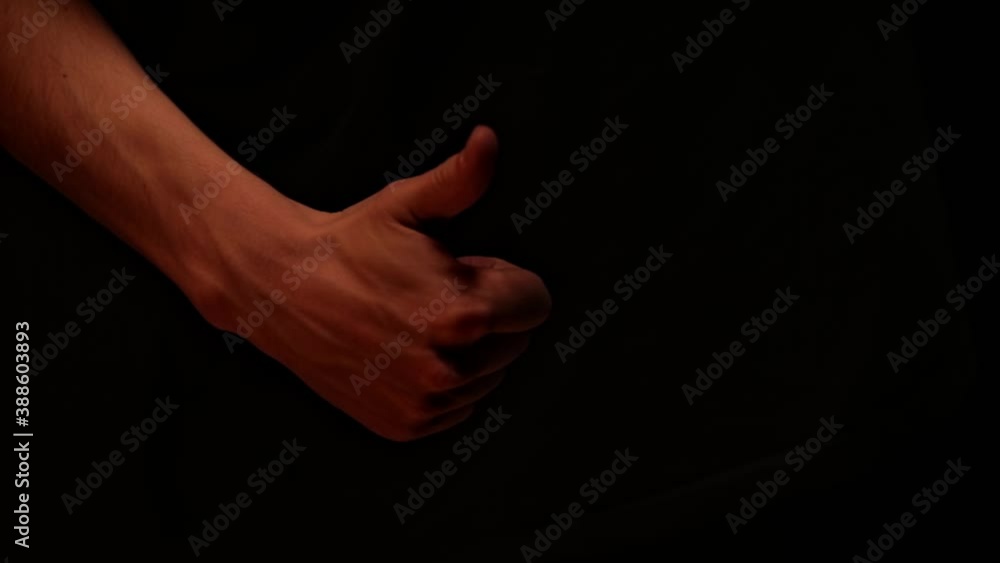 The man his hand showing the thumb raised up. A man's hand shows a like sign and shakes her hand on dark background The man his hand showing the thumb raised up. A man's hand shows a like sign and shakes her hand on dark background