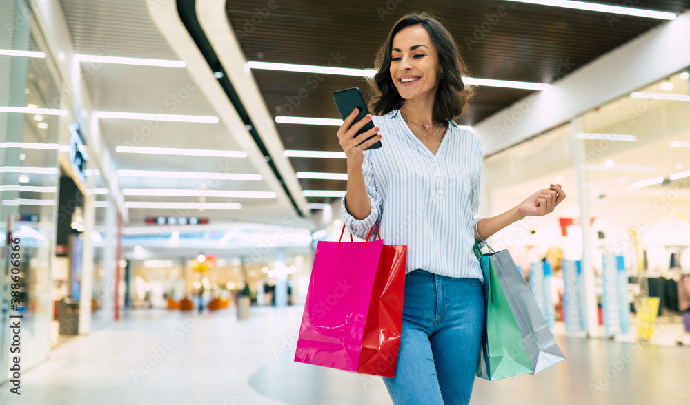 Modern gorgeous young smiling woman in trendy stylish clothes with bright colorful shopping bags is using her smart phone while walking in the mall Modern gorgeous young smiling woman in trendy stylish clothes with bright colorful shopping bags is using her smart phone while walking in the mall
