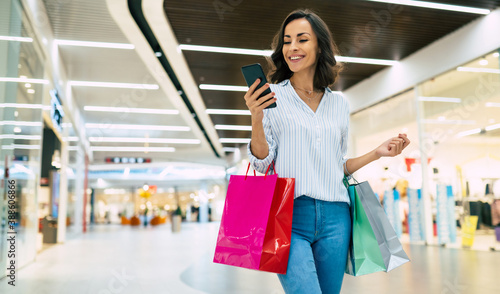 Modern gorgeous young smiling woman in trendy stylish clothes with bright colorful shopping bags is using her smart phone while walking in the mall