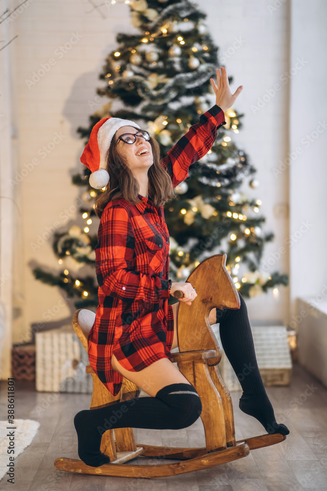 Pretty woman in shirt and socks have fun riding on the wooden swing horse toy, new year tree behind.