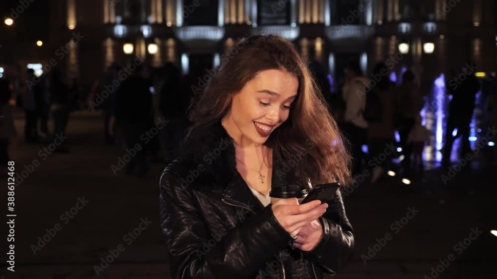 Cheerful girl holding a phone and coffee in her hands and smiling on a background of a beautiful purple fountain at night. Cheerful girl holding a phone and coffee in her hands and smiling on a background of a beautiful purple fountain at night.