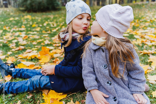 фотографія Little cute stylish long hair girls, kissing sisters in autumn coats sitting in multi-colored yellow red maple leaves, foliage