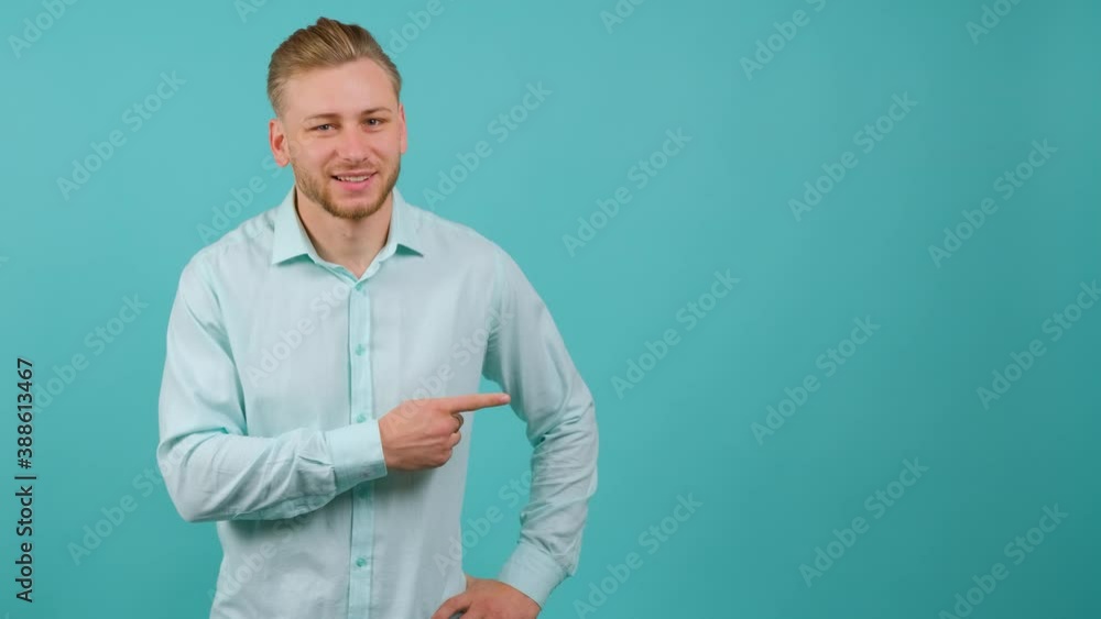 A confident looking young bearded man is pointing to the left and doing thumb up gesture, standing isolated over turquoise background. A confident looking young bearded man is pointing to the left and doing thumb up gesture, standing isolated over turquoise background.