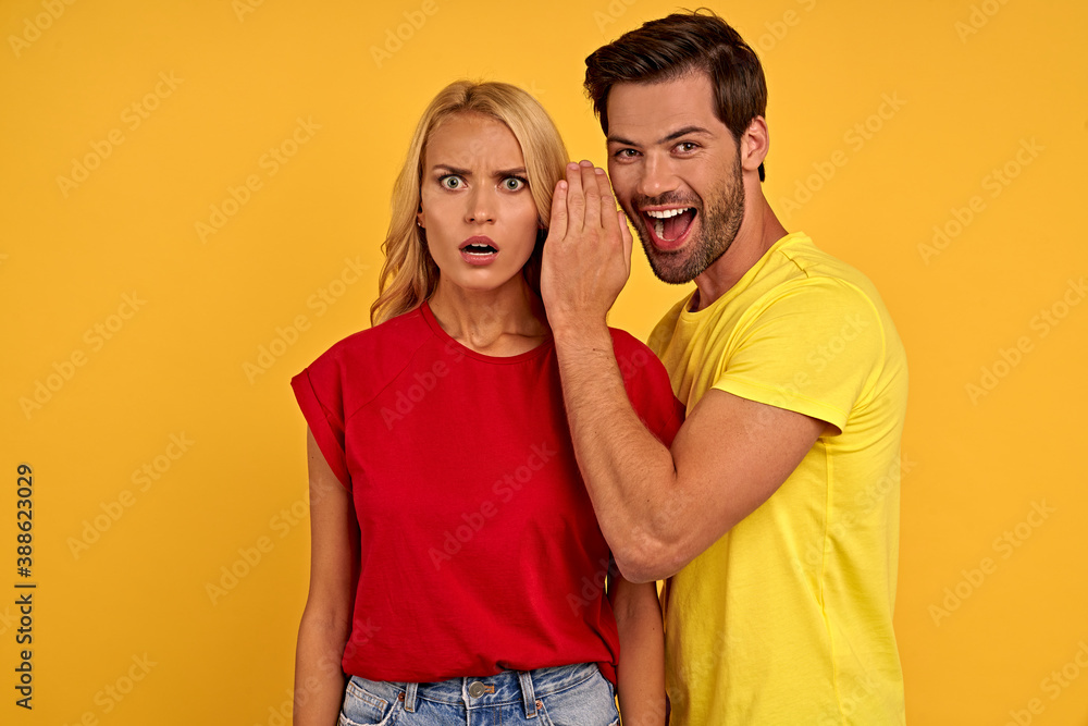Naklejka premium Excited young couple friends guy girl in casual clothes isolated on yellow background. People lifestyle concept. Mock up copy space. Whispers gossip and tells secret behind hand, doing winner gesture
