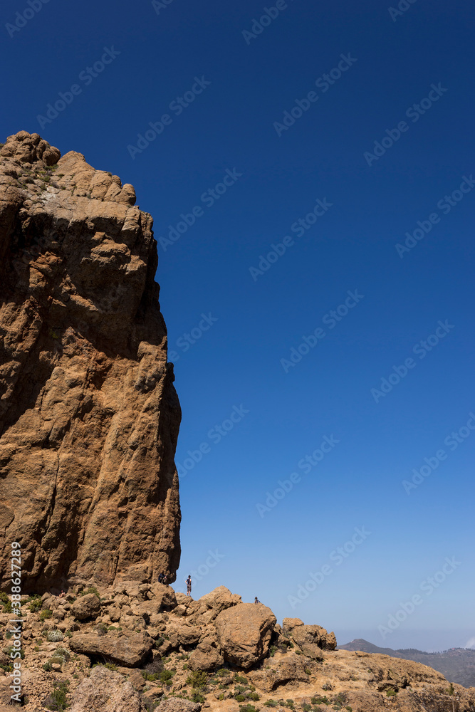Hikers Seem Dwarfed Next To The Enormous Volcanic Rock Roque Nublo (Rock in the Clouds), One Of The Highest Peaks On The Island Of Gran Canaria, Canary Islands, Spain. Vertical Establishing Shot