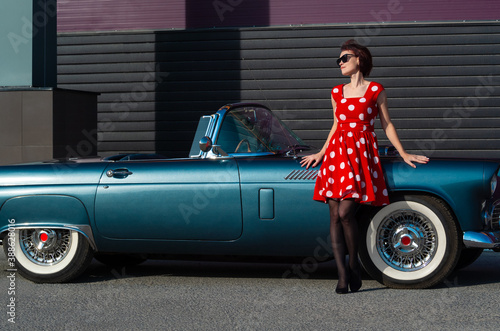 A young lady in a red polka dot dress and sunglasses stands near a vintage car.