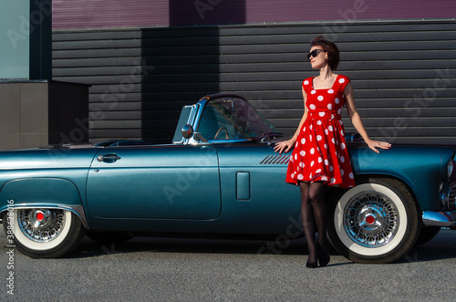 Bilde på lerret A young lady in a red polka dot dress and sunglasses stands near a vintage car