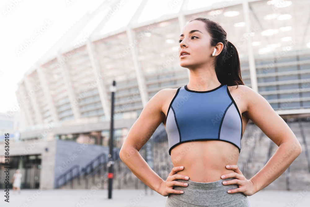 Confident fit caucasian female runner standing against big modern stadium in sporty top with earbuds