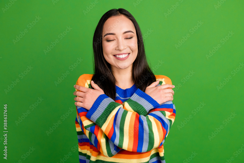 Close-up portrait of lovely dreamy cheerful girl wearing striped warm pullover embracing herself isolated over vibrant green color background Close-up portrait of lovely dreamy cheerful girl wearing striped warm pullover embracing herself isolated over vibrant green color background