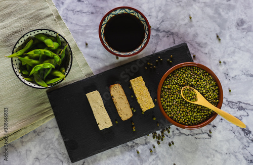 Top view of different products derived from soy. Edamame beans, soy sauce, tofu, tempeh, and soybeans. Food on a tablecloth on a marble countertop.