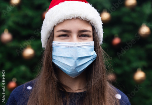 Close up Portrait of woman wearing a santa claus hat and medical mask with emotion. Against the background of a Christmas tree. Coronavirus pandemic © lunarts_studio
