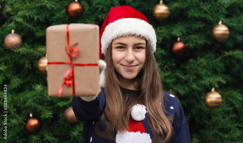 A Christmas gift. A young girl wearing a santa claus hat gives a gift made of craft paper with a red ribbon. Against the background of a Christmas tree A Christmas gift. A young girl wearing a santa claus hat gives a gift made of craft paper with a red ribbon. Against the background of a Christmas tree