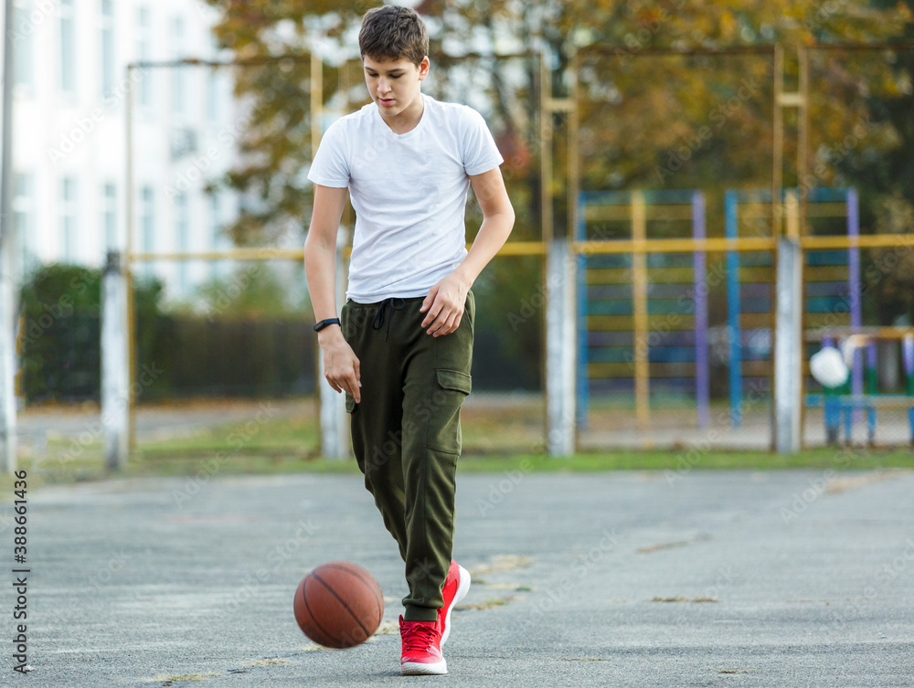 Cute young boy plays basketball on street playground. Teenager in white t shirt with orange basketball ball outside. Hobby, active lifestyle, sport activity for kids.