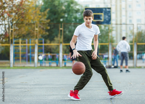 Cute young boy plays basketball on street playground. Teenager in white t shirt with orange basketball ball outside. Hobby, active lifestyle, sport activity for kids.