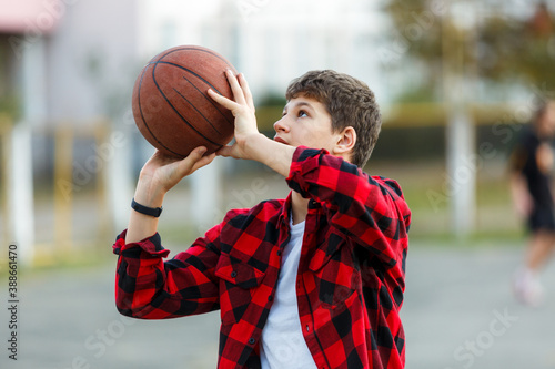Cute young boy plays basketball on street playground. Teenager in red check flannel shirt with orange basketball ball outside. Hobby, active lifestyle, sport activity for kids. © Natali