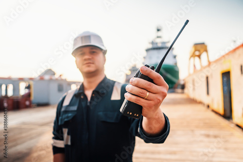 Marine Deck Officer or Chief mate on deck of offshore vessel or ship , wearing PPE personal protective equipment - helmet, coverall. He holds VHF walkie-talkie radio in hands. photo