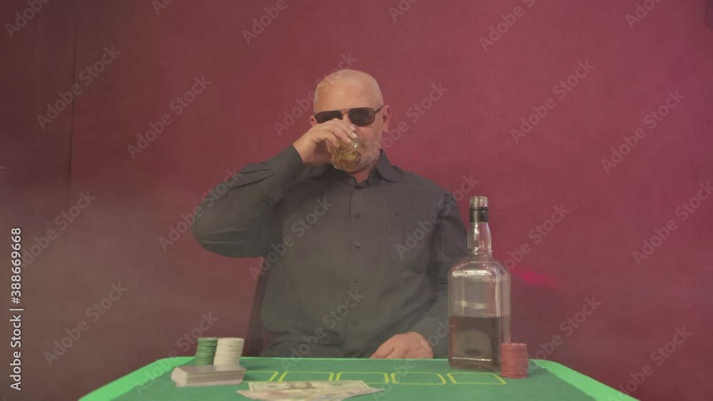 A man in a casino at a gambling table raises a glass of whiskey. Green laser lights and smoke in the background. There are chips, cards and dollars. The player is wearing black glasses. UHD. A man in a casino at a gambling table raises a glass of whiskey. Green laser lights and smoke in the background. There are chips, cards and dollars. The player is wearing black glasses. UHD.