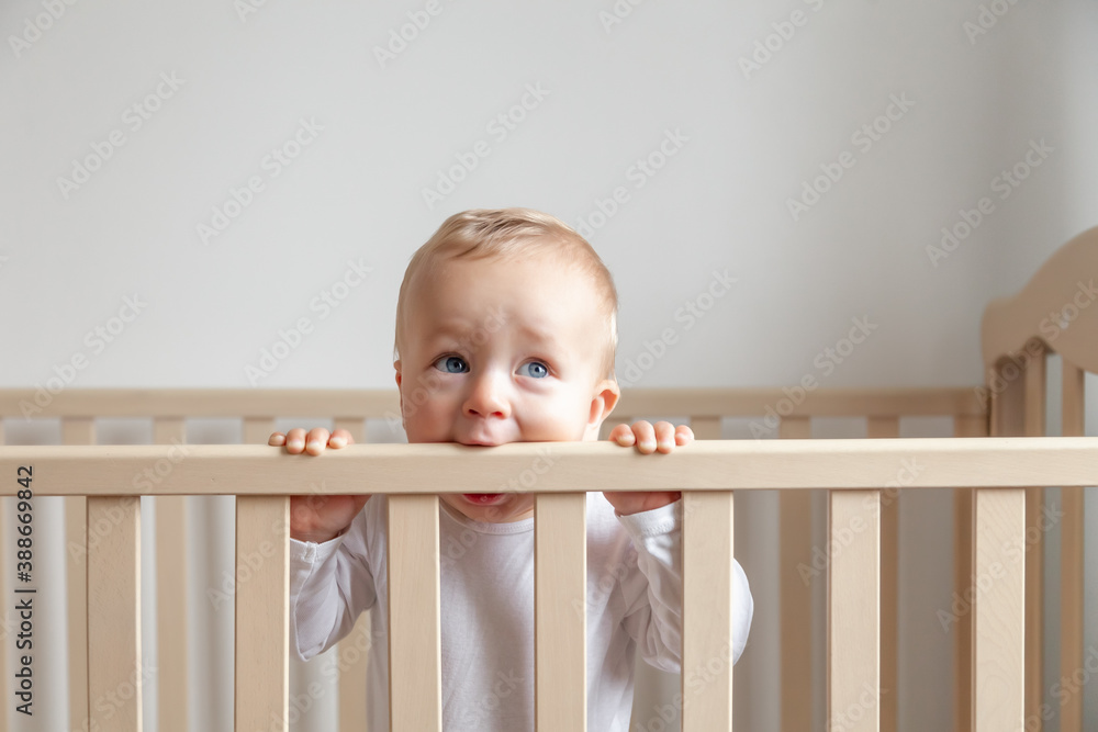 Blond cute little baby biting wooden bed headboard - obrazy, fototapety, plakaty 