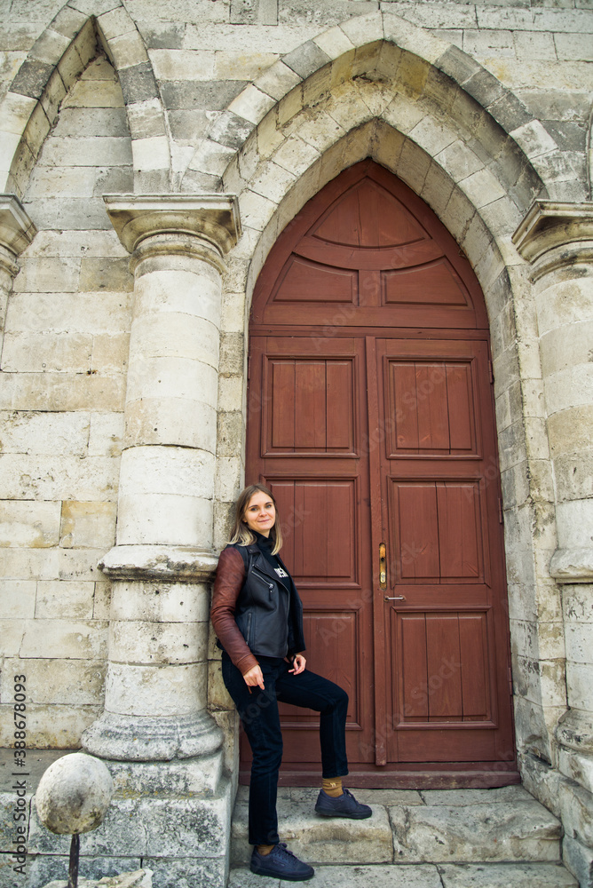 young woman in a leather jacket near the entrance to the ancient wooden door of a gothic temple