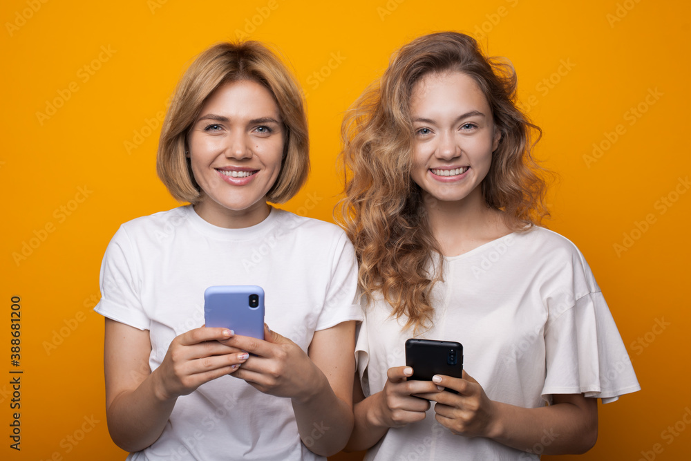 Two caucasian blonde sisters are chatting with phones while smiling at camera on a yellow studio wall Two caucasian blonde sisters are chatting with phones while smiling at camera on a yellow studio wall