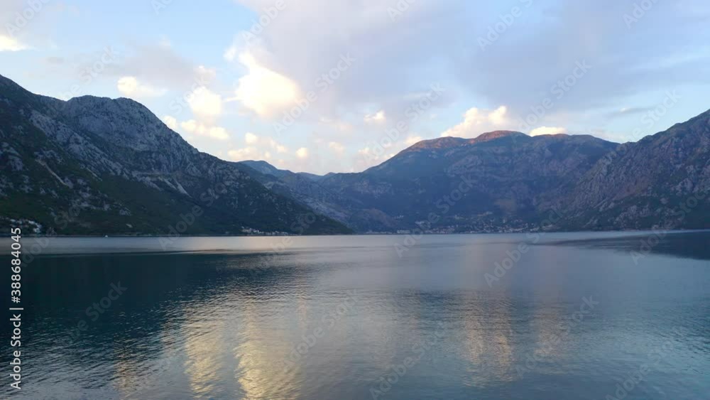 The bay of Kotor in Montenegro, with its still waters, surrounded by high mountain range, reflected in the water, white clouds above the horizon, late afternoon sunlight shining over the peaks. The bay of Kotor in Montenegro, with its still waters, surrounded by high mountain range, reflected in the water, white clouds above the horizon, late afternoon sunlight shining over the peaks.