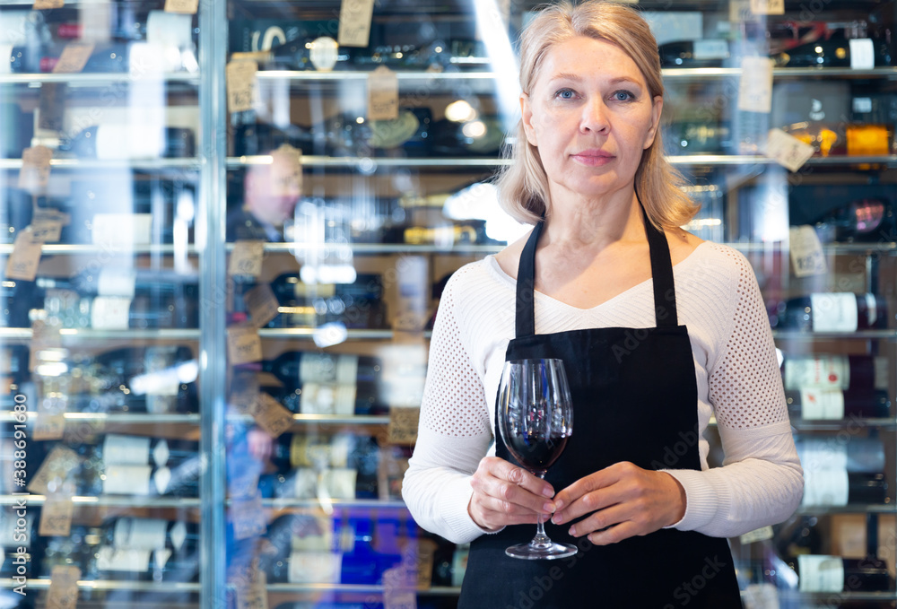 Confident mature female winemaker inviting to wine house, offering glass of wine for tasting..