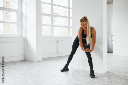 A beautiful athletic model with a slim body in trendy sportswear doing a leg stretch in a bright room. blonde in a black suit warms up. healthy lifestyle