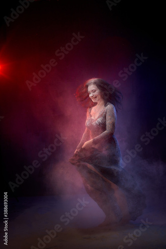 Girl in black dress posing in dark studo during photoshoot with flour or dust and light. Dangerous witch during struggle between good and evil