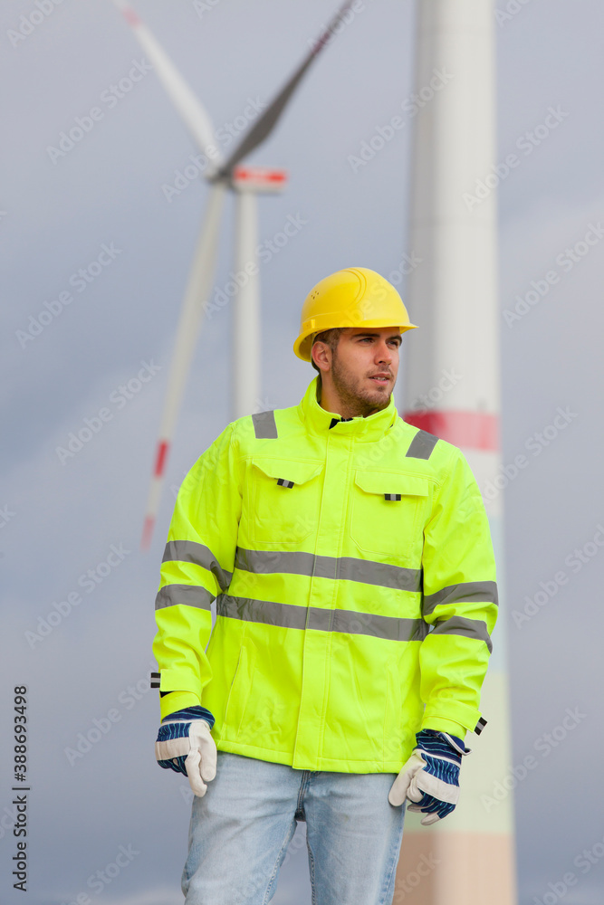 Smiling young engineer with hard hat and protective clothing in front of blurred wind turbines Smiling young engineer with hard hat and protective clothing in front of blurred wind turbines