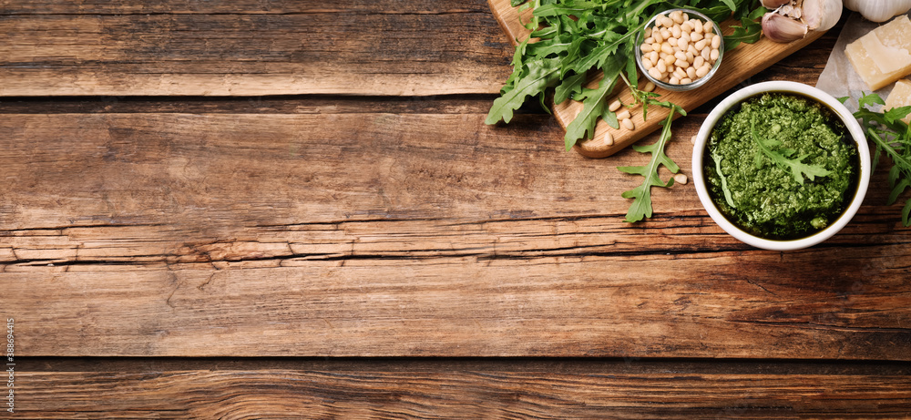 Bowl of tasty arugula pesto and ingredients on wooden table, flat lay with space for text. Banner design Bowl of tasty arugula pesto and ingredients on wooden table, flat lay with space for text. Banner design