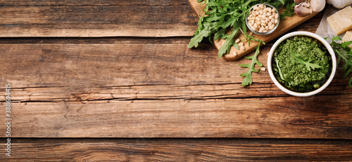 Bowl of tasty arugula pesto and ingredients on wooden table, flat lay with space for text. Banner design