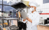 cooking, culinary and health concept - female chef in toque wearing face protective medical mask for protection from virus disease with tablet pc computer over kebab shop kitchen background