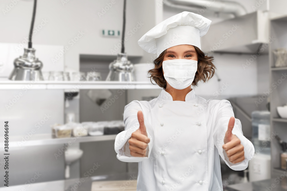 cooking, culinary and health concept - female chef in toque wearing face protective medical mask for protection from virus disease showing thumbs up over restaurant kitchen background