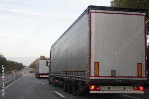 White van semi trucks convoy drive on right lane of empty two way suburban asphalted highway road at summer day, back side view – international logistics, cargo transportation, trucking industry