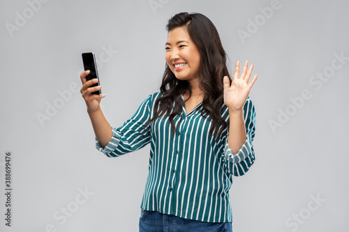 technology, blogging and people concept - happy asian woman taking selfie with smartphone or having video call and waving hand over grey background