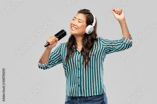 people, leisure and music concept - happy smiling young asian woman in headphones with hairbrush singing over grey background