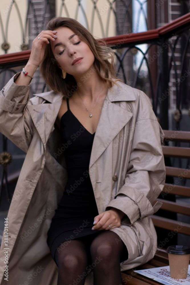 Fototapeta premium attractive slim young brunette in fashionable trench coat and black dress sitting on a bench with a newspaper. autumn, yellow leaves.