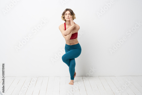 sports woman in a bright room doing yoga in full growth blue leggings and a red tank top