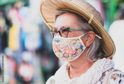 Side view of a smiling senior woman face with glasses and scarf wearing a medical mask due to coronavirus enjoying  the flea market with an explorer's hat