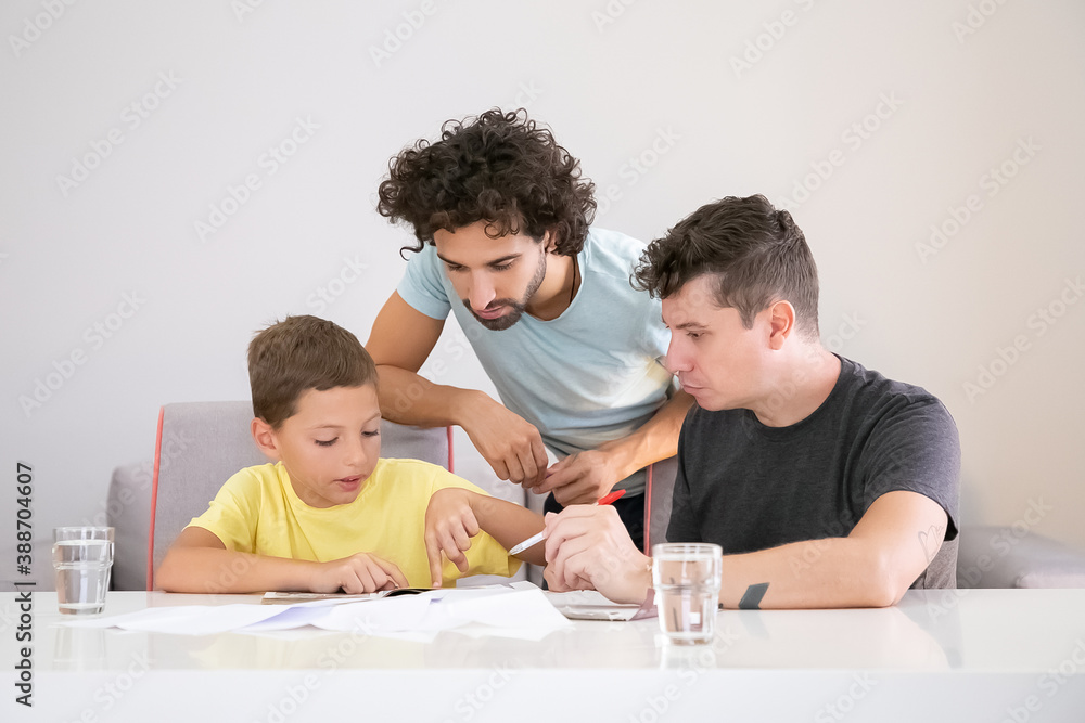 Two dads helping focused boy with school home task, sitting at table with papers, reading textbook together, pointing finger at page. Family and parenthood concept Two dads helping focused boy with school home task, sitting at table with papers, reading textbook together, pointing finger at page. Family and parenthood concept
