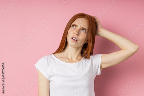 Pensive young caucasian ginger female with freckled skin long hair wearing white t shirt looking up with puzzled expression, touching lips with forefinger, thinking isolated blue background.Copy space