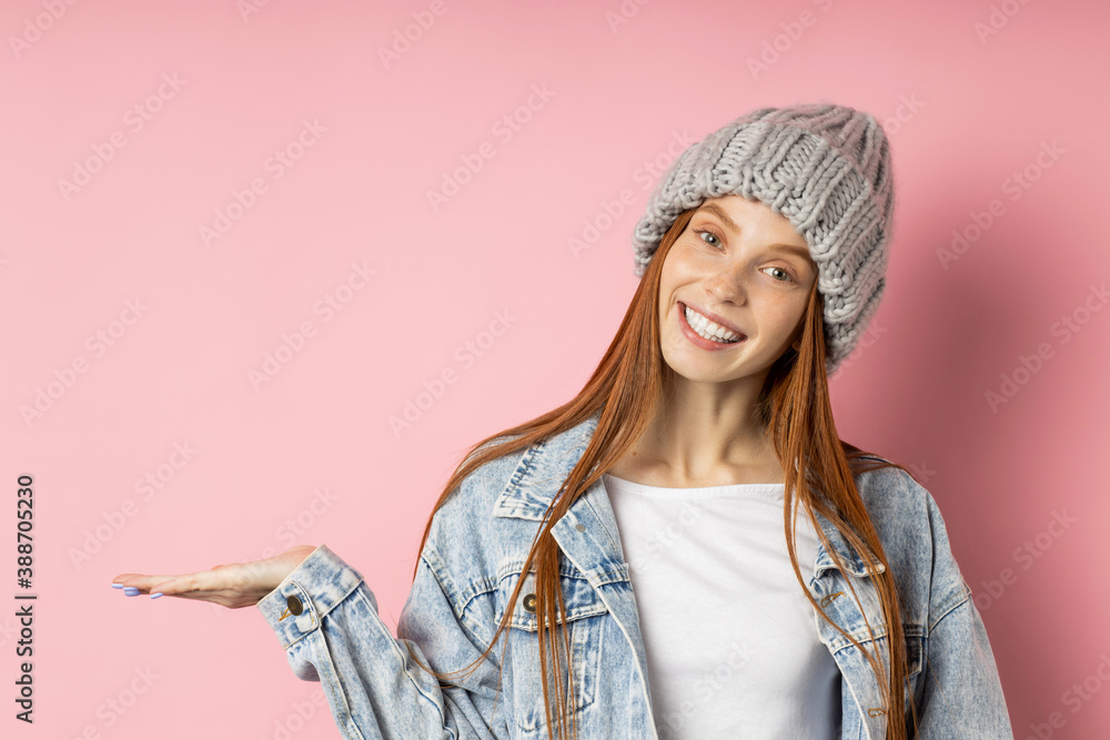Satisfied european girl with red hair in oversized denim jacket, gray winter hat, keeping palm raised, holding copy space, advertising your product isolated pink background. People,promotion concept. Satisfied european girl with red hair in oversized denim jacket, gray winter hat, keeping palm raised, holding copy space, advertising your product isolated pink background. People,promotion concept.