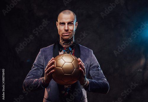 Smirking guy in dark custom suit with tattoo and short haircut poses with golden soccer ball in dark background.