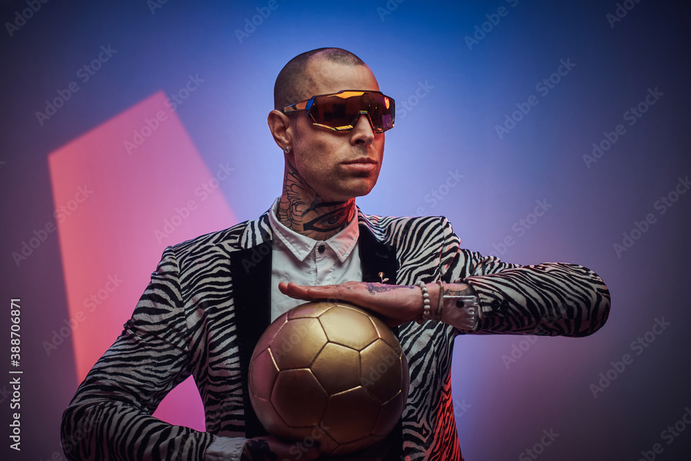 Plakat Handsome guy dressed in custom suit with short haircut and eyewears poses with golden soccer ball in abstract background.