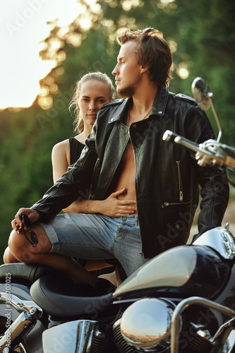 young couple of bikers closely looking at each other on black motorcycle on road near green forest