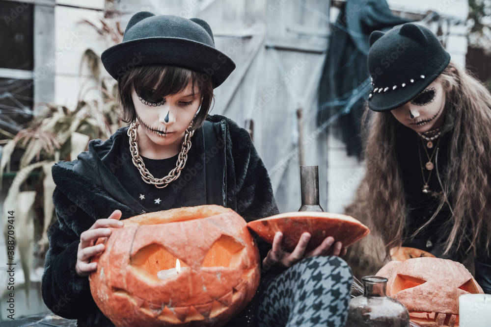Scary little girls, sisters celebrating halloween. Pumpkin jack-o-lantern.Terrifying black face makeup,witch costumes,stylish image.Horror,fun at children's party in barn on street.Hat,fur coat,chain