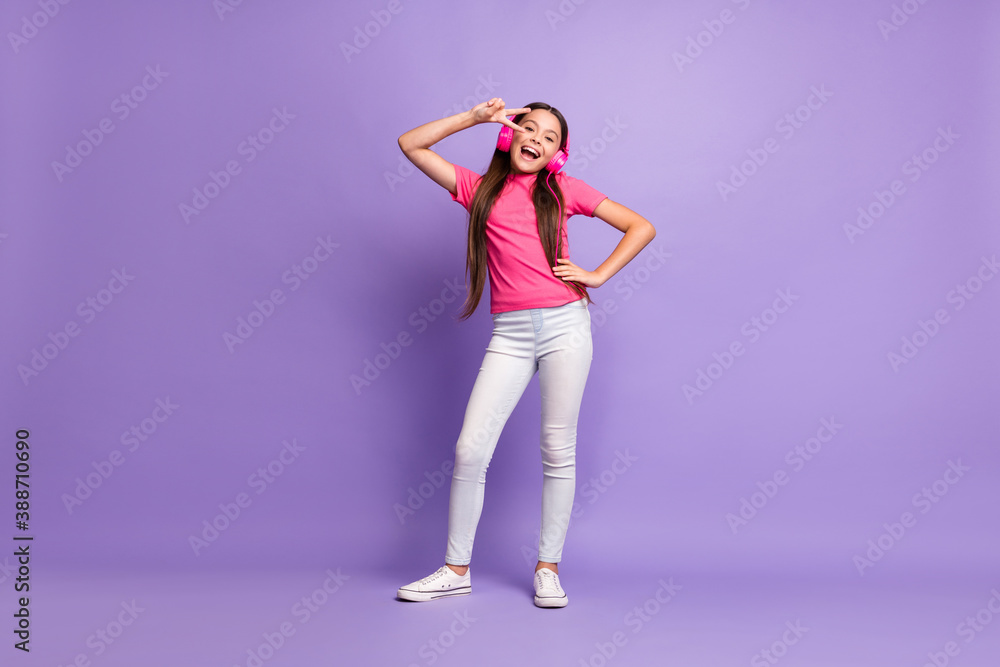 Full length body size photo of positive little girl smiling listening to music showing gesture peace v-sign isolated on purple color background Full length body size photo of positive little girl smiling listening to music showing gesture peace v-sign isolated on purple color background