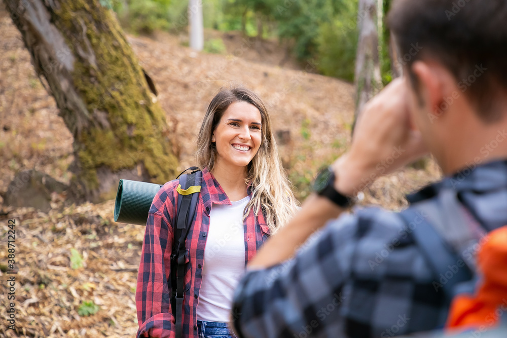 Happy lady posing and smiling on road in forest. Unrecognizable man taking photo of his girlfriend. Tourists hiking together in woods and having fun. Tourism, adventure and summer vacation concept
