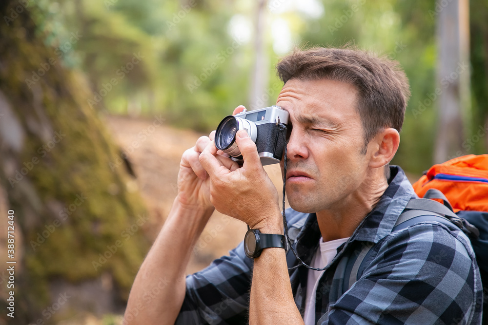 Focused man shooting landshate and walking in forest. Caucasian traveler exploring nature, holding camera, taking photo and carrying backpack. Tourism, adventure and summer vacation concept Focused man shooting landshate and walking in forest. Caucasian traveler exploring nature, holding camera, taking photo and carrying backpack. Tourism, adventure and summer vacation concept