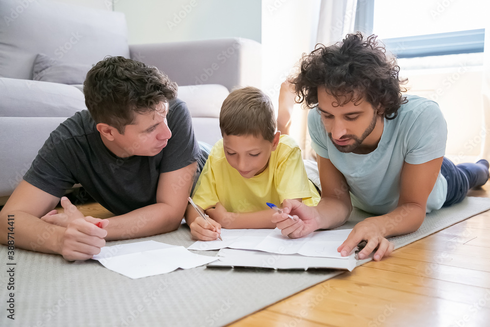 Couple of homosexual parents helping focused boy with school home task, lying on floor at home, writing or drawing in papers. Family and gay parents concept Couple of homosexual parents helping focused boy with school home task, lying on floor at home, writing or drawing in papers. Family and gay parents concept